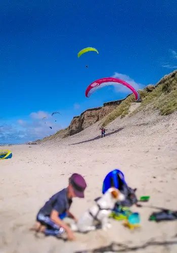Gleitschirmpiloten fliegen über den Dünen von Lyngby Beach, während ein Kind mit Hund im Vordergrund spielt.