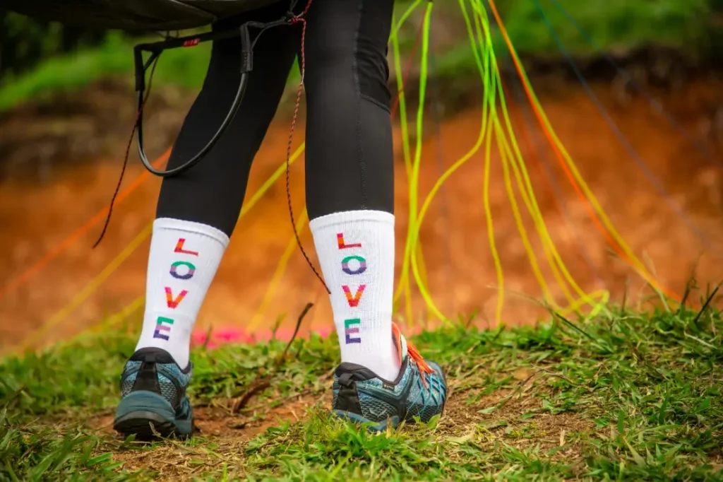 Girl's legs on a green take-off field, a paraglider in the background, white socks with the word "LOVE" in colorful letters.