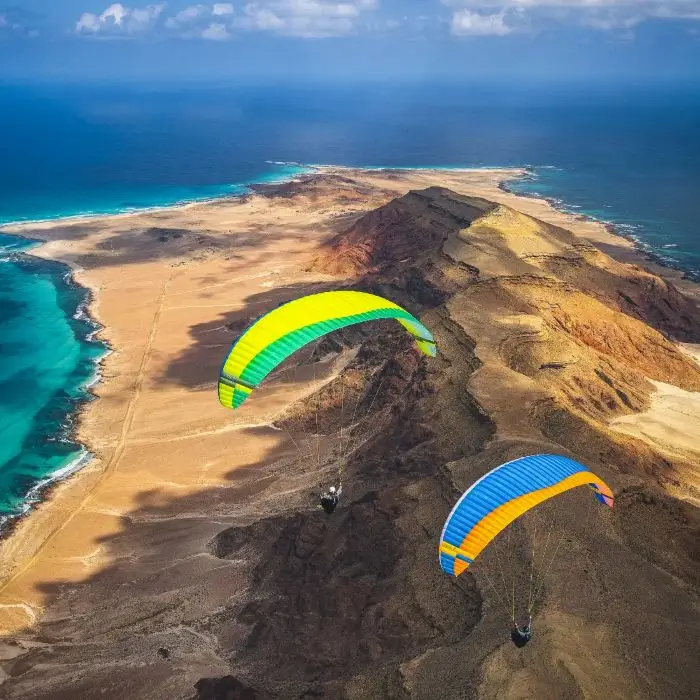 Mehrere Gleitschirme fliegen über goldene Sanddünen mit atemberaubendem Blick aufs blaue Meer – das perfekte Abenteuer bei einer Flugreise.